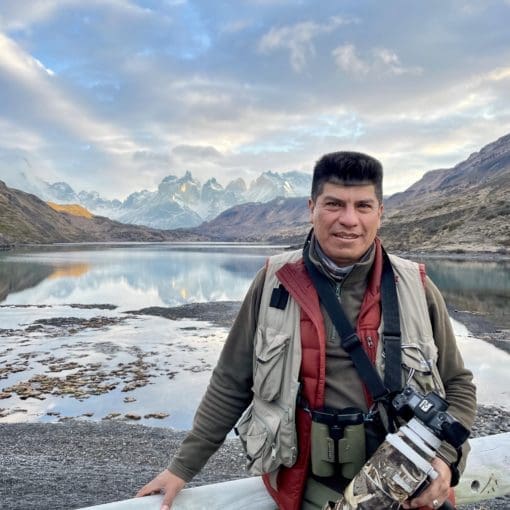 A man stands in outdoor clothes for cold weather in front of a gorgeous view of mountains and sky reflecting in a lake