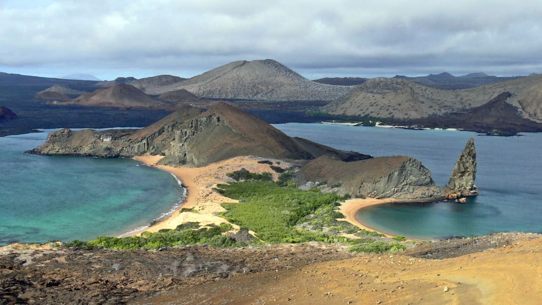 View of Santiago Island from Bartolome Island includes several volcanic domes, stunning blue waters, and green vegetation against golden sand