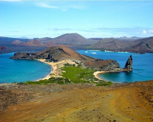 View of Santiago Island in the distance and Bartolome in the foreground. The two dark volcanic landscapes are divided by brilliant blue waters dotted with yachts 