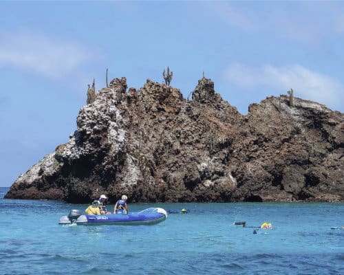 A tour group snorkels in brilliant blue water next to a sparse, rocky outcropping with a nearby dinghy